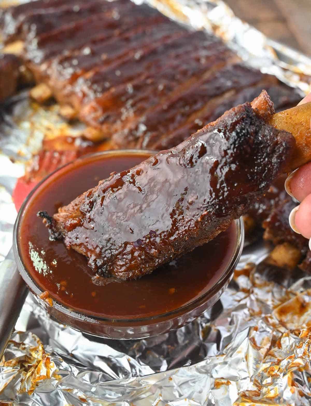 A pork rib being dipped in a side of bbq sauce.
