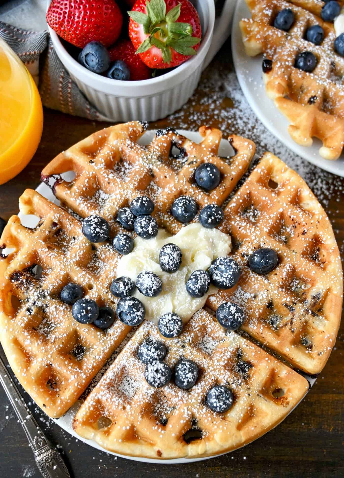 blueberry waffle on a plate with fresh blueberries, butter and powdered sugar on top.