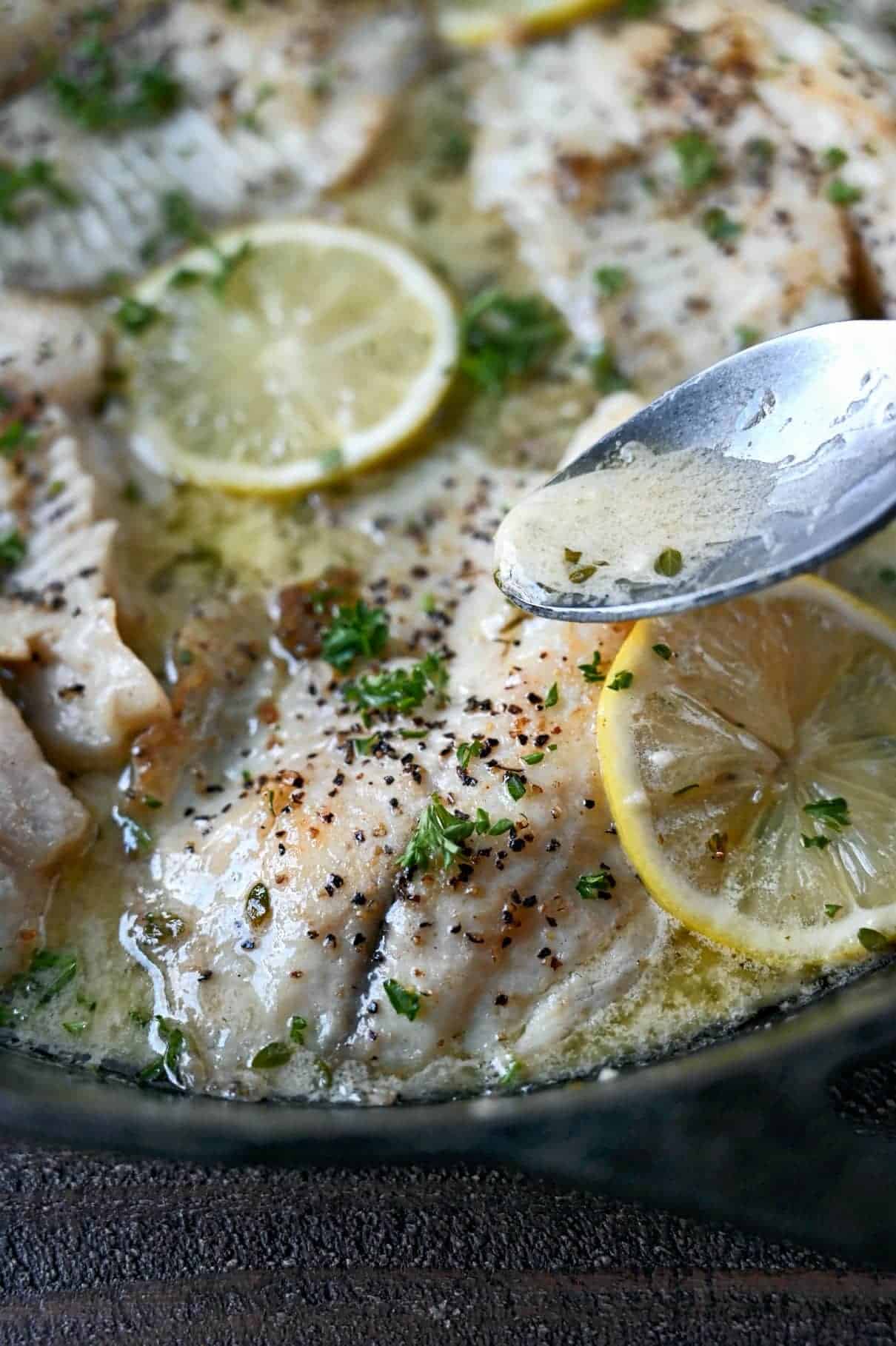 Tilapia in a skillet with fresh lemon slices and lemon sauce being poured on top with a spoon.