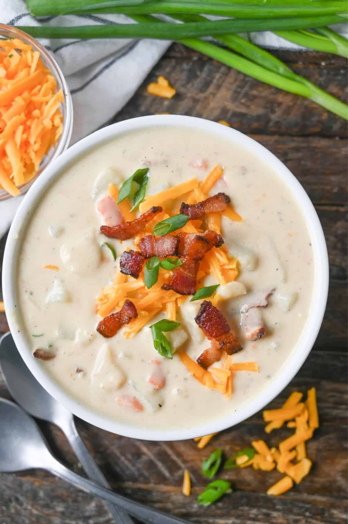 Loaded potato soup in a white bowl with cheese, bacon and green onions on top. And a small bowl of grated cheddar cheese on the side.