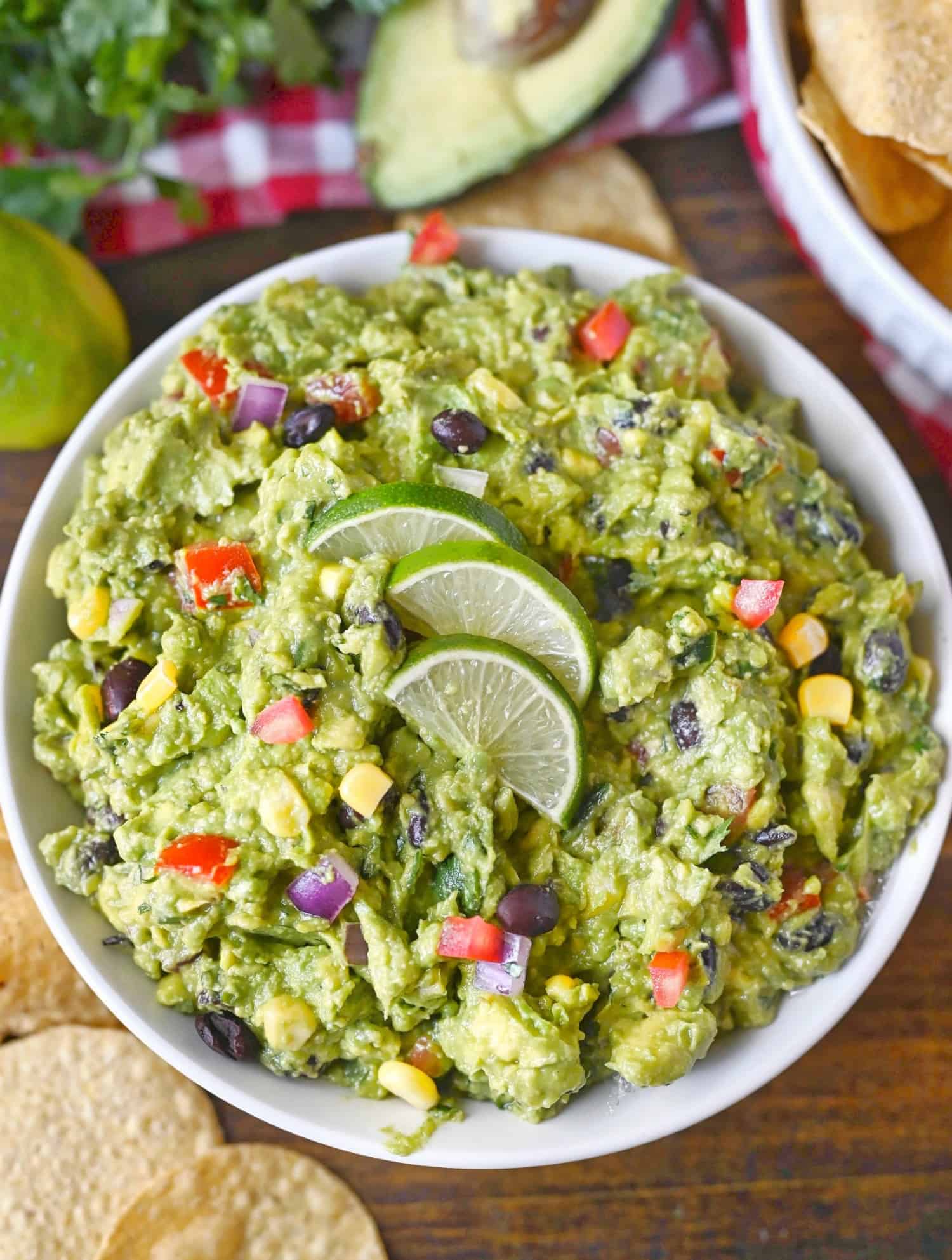 Cowboy guacamole in a white bowl with sliced limes on top. A side of chips and some limes and avocados on the side.