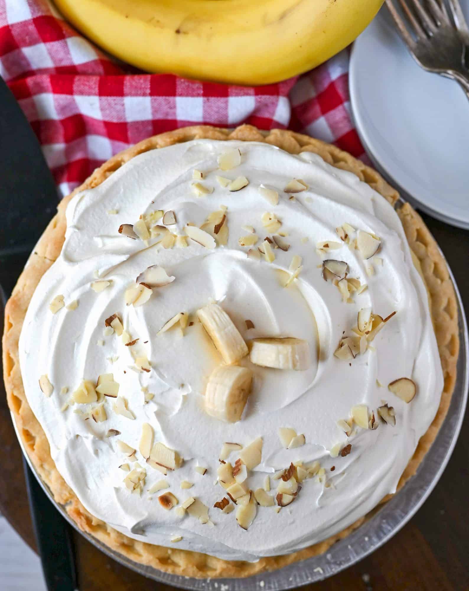 Banana crem pie with banana bunch on the side on top of a red chechered linen. Small whites plate and forks on the side.