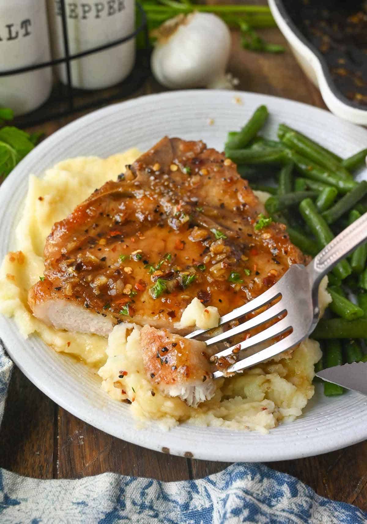 Brown sugar garlic pork chop with a piece cut on a plate with mashed potatoes and green beans.