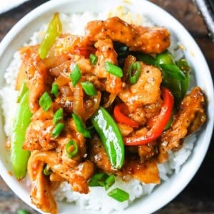 Sweet n spicy chicken stir fry on top of white rice in a white bowl with green onions sprinkled on top.