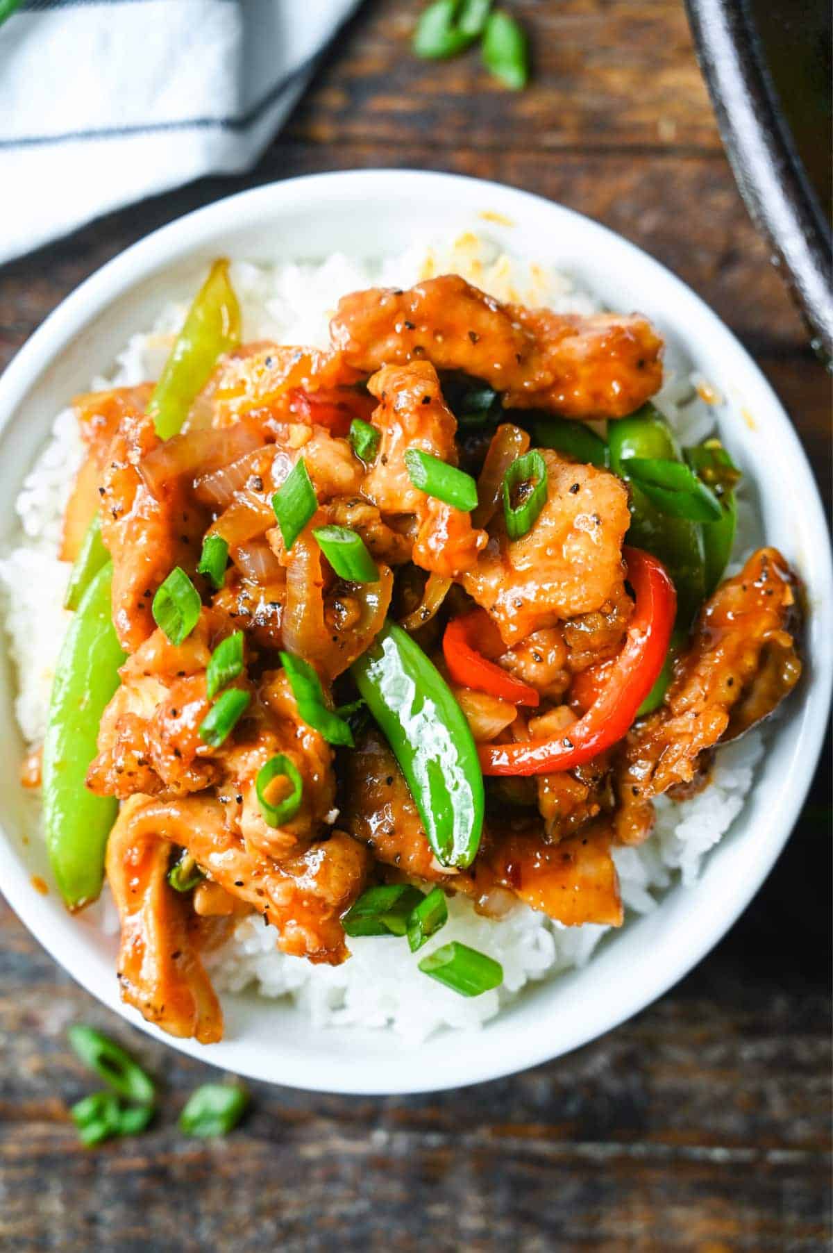 Sweet n spicy chicken stir fry on top of white rice in a white bowl with green onions sprinkled on top.