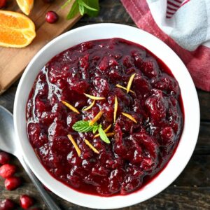 Homemade cranberry sauce in a white bowl with orange peel shavings on top and a sprig of mint.