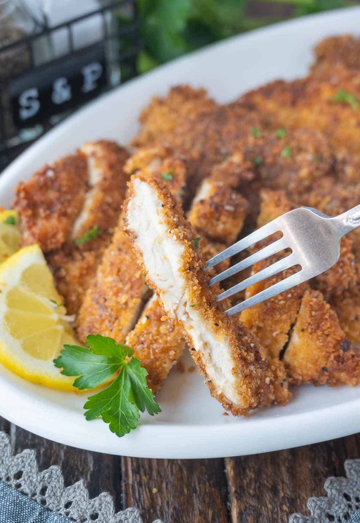 Crispy chicken cutlets cut into slices on a white plate with a lemon slice.