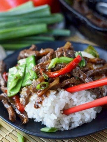 Beef stir fry with white rice on a black plate and red chopsticks