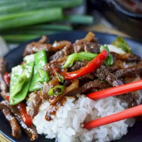 Beef stir fry with white rice on a black plate and red chopsticks