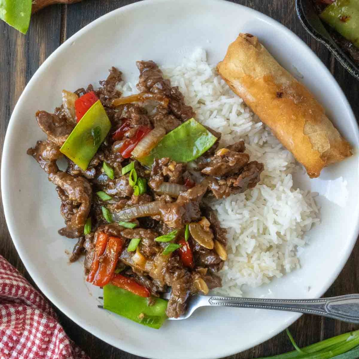 Beef stir fry on a white plate with rice and egg roll.