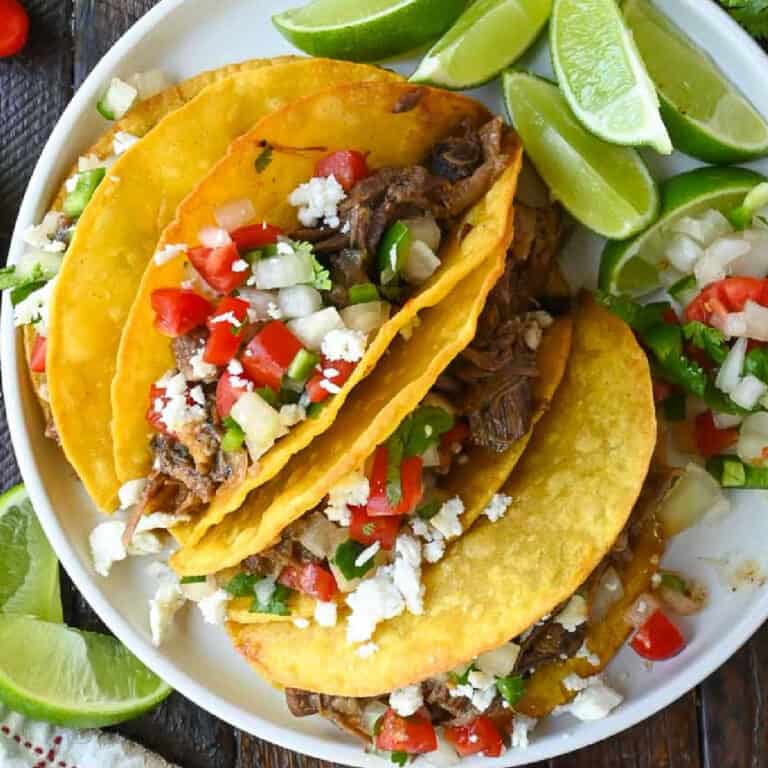 Four beef short rib tacos on a white plate with sliced limes on the side.