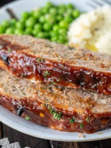 Two slices of meatloaf on a plate with potatoes and peas on a plate.