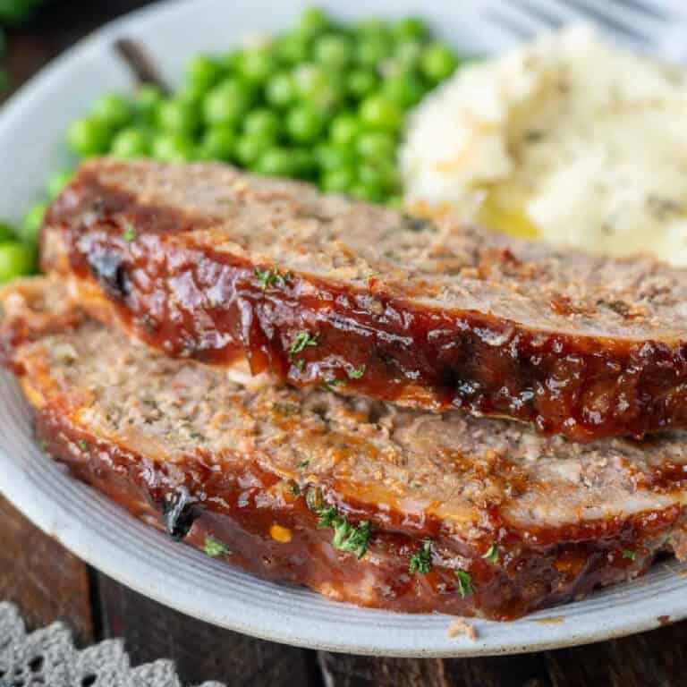 Two slices of meatloaf on a plate with potatoes and peas on a plate.
