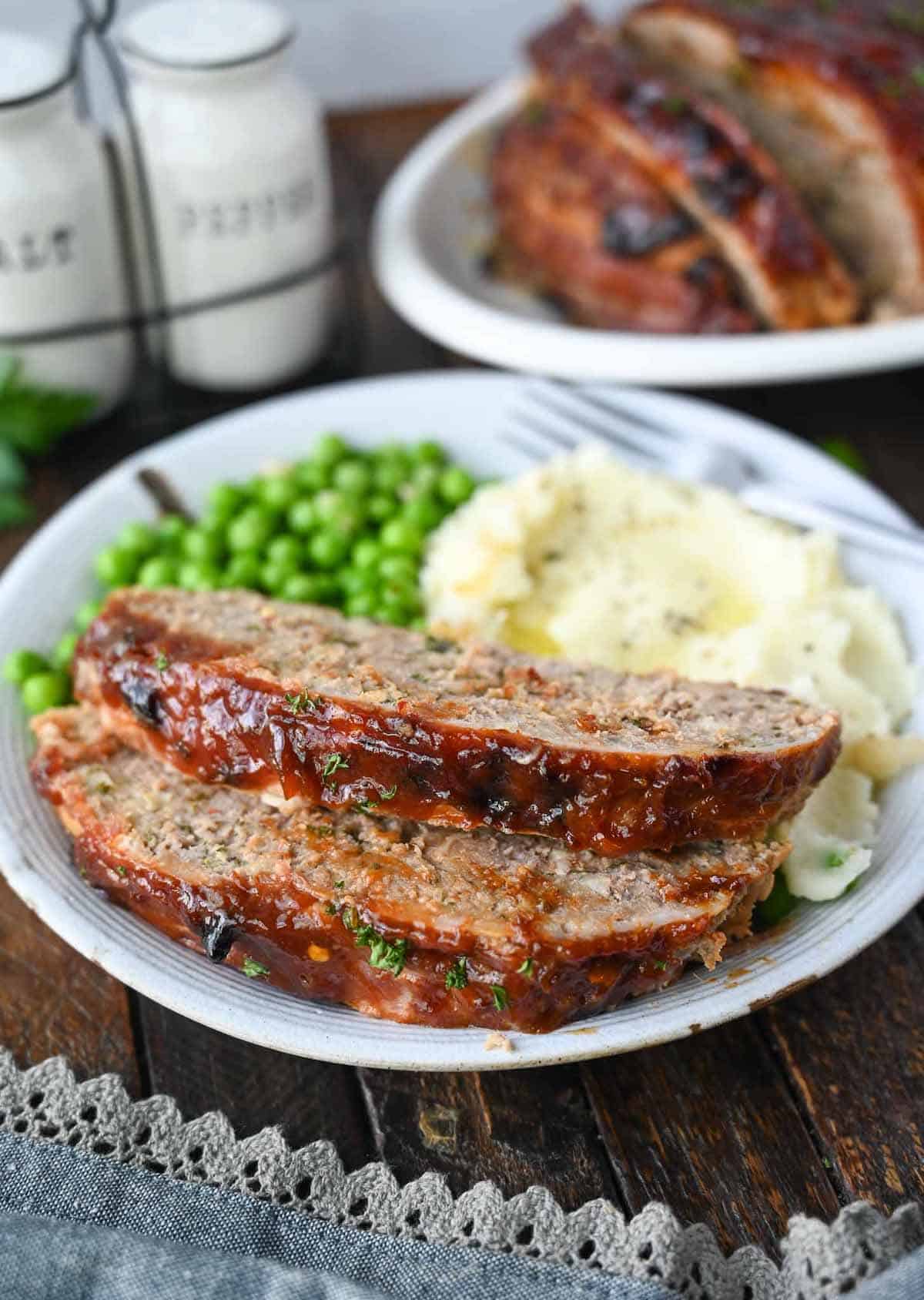 Two slices of meatloaf on a plate with potatoes and peas on a plate.