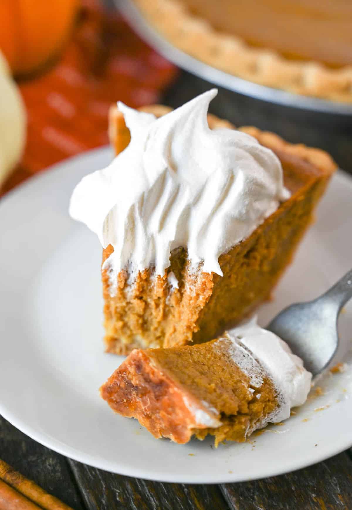 A bite of pumpkin pie on a fork laying next to a piece of pie.