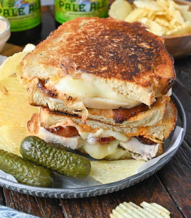 Turkey grilled cheese on a plate with pickles and chips.