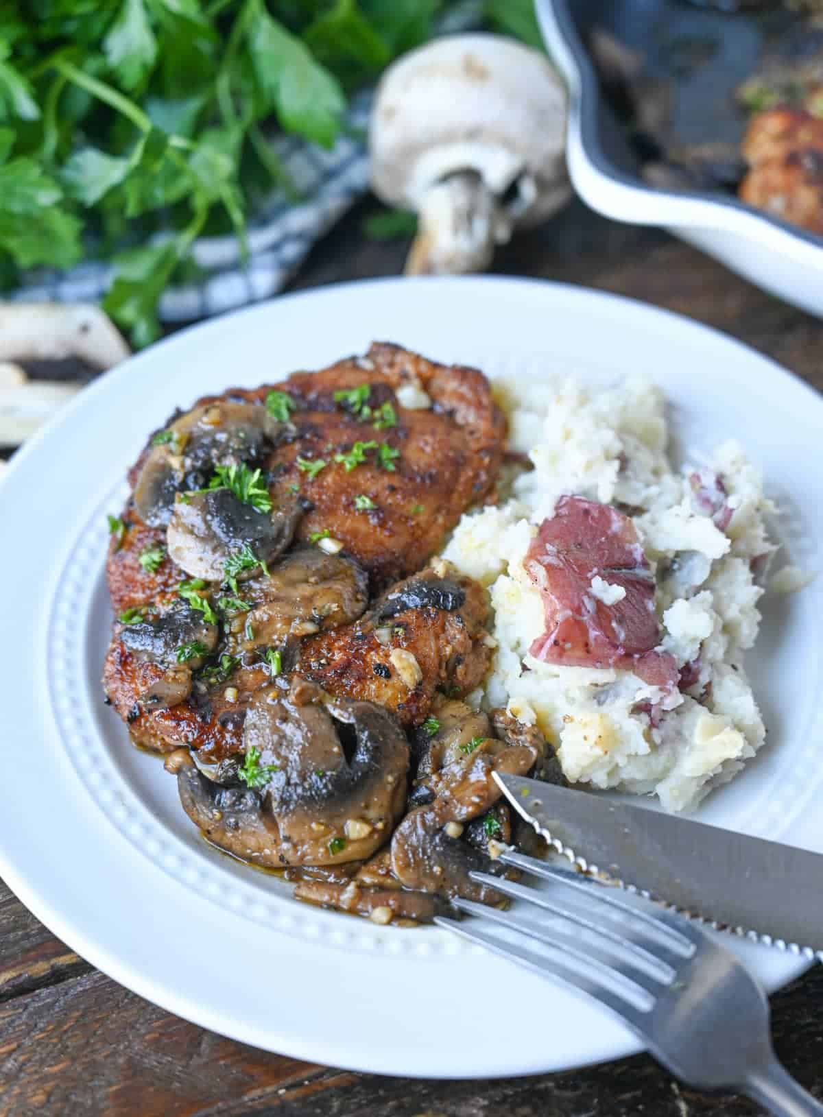 Chicken thigh with garlic mushrooms on top with a side of red mashed potatoes on a white plate.