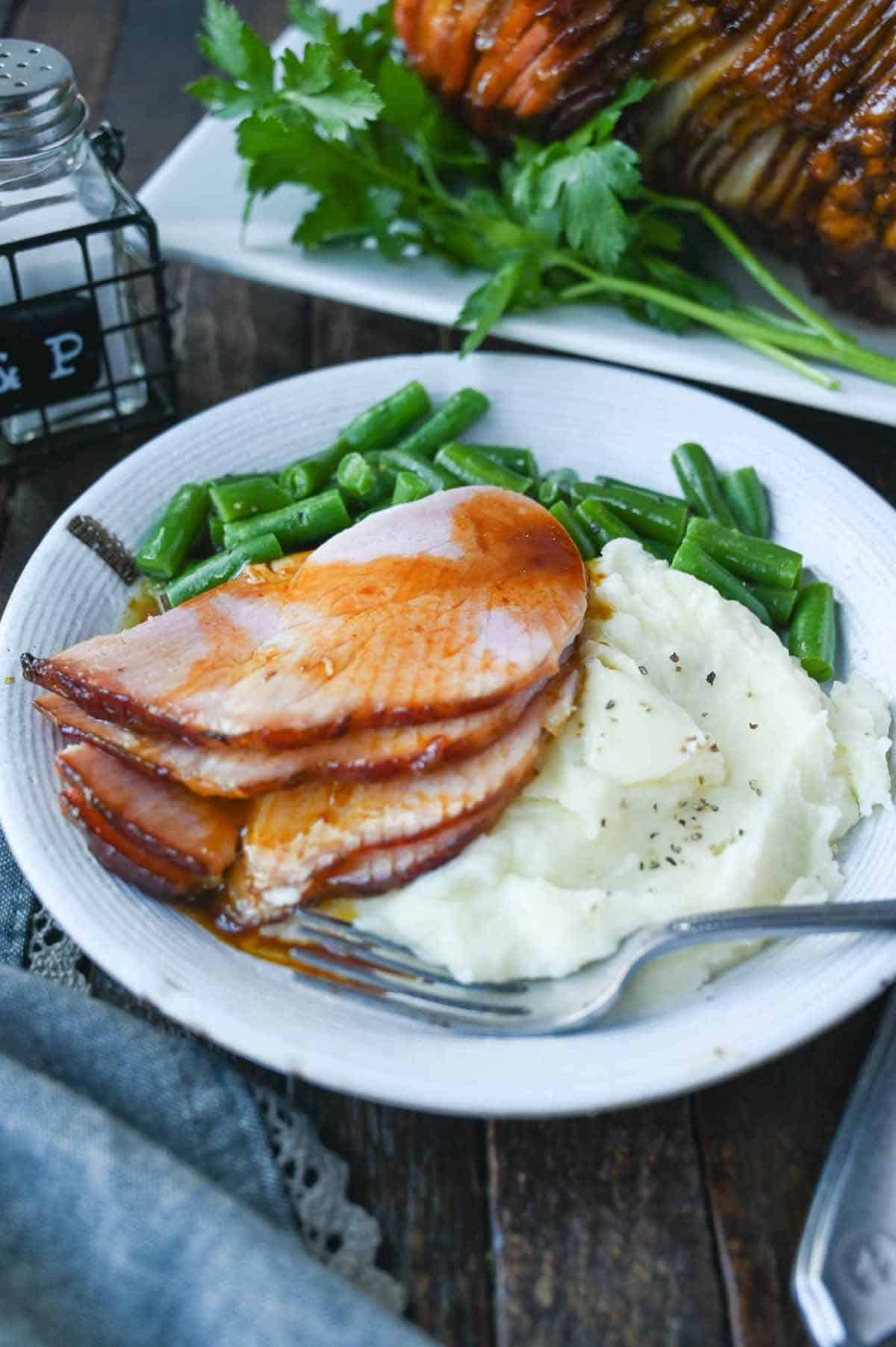 Spiral honey ham on a white plate with mashed potatoes and green beans.