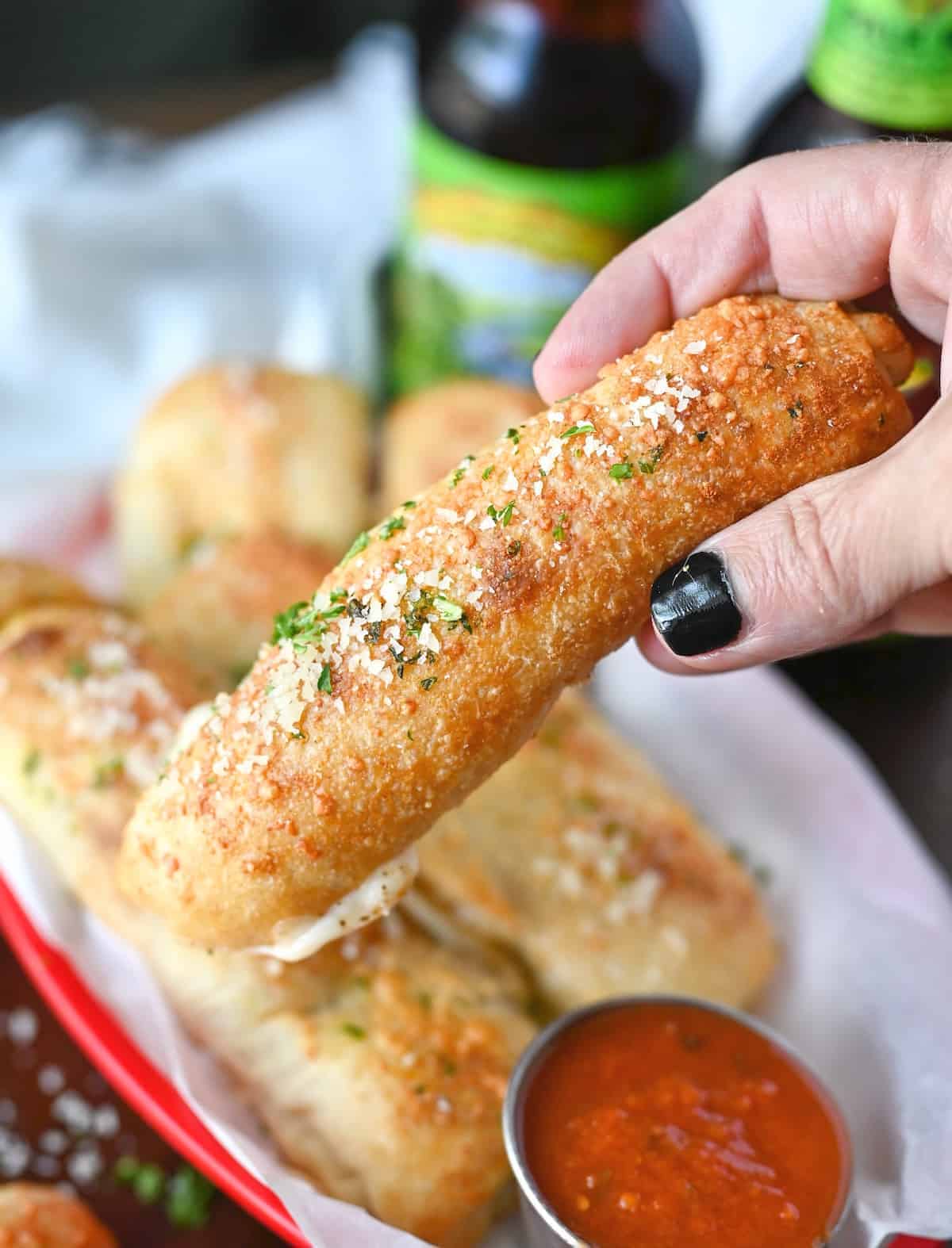 One pizza stick being picked up from a red serving basket with a side of pizza sauce.