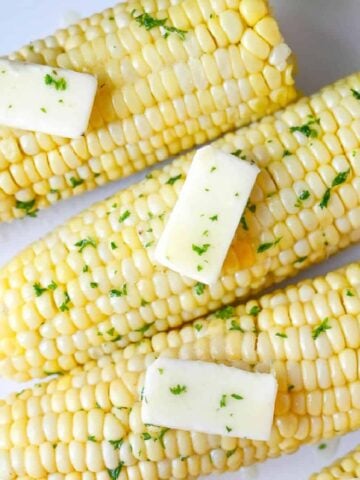 Four ears of corn on a white plate with butter.