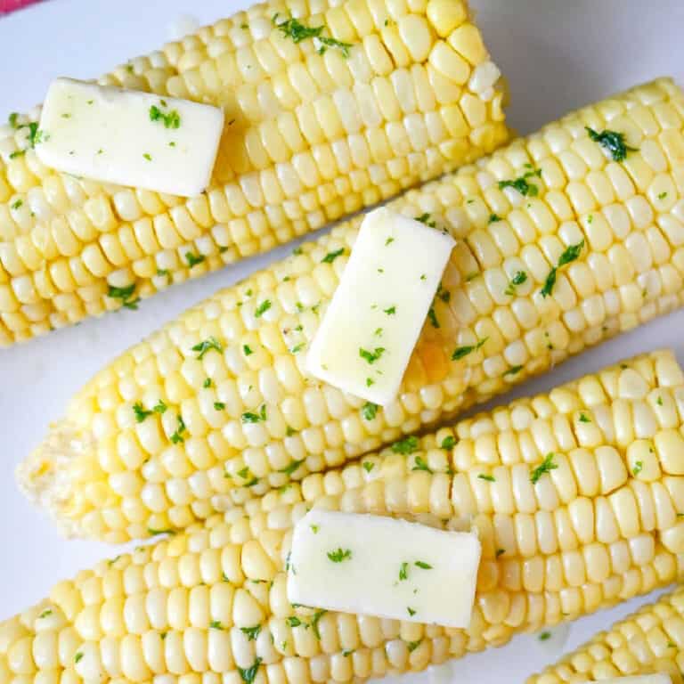 Four ears of corn on a white plate with butter.