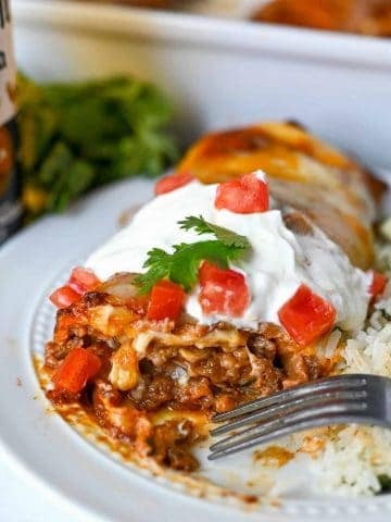 beef burrito on a white plate with sourcream on top and diced tomatoes. the end cut off and gooey, cheesy beef is oozing out.