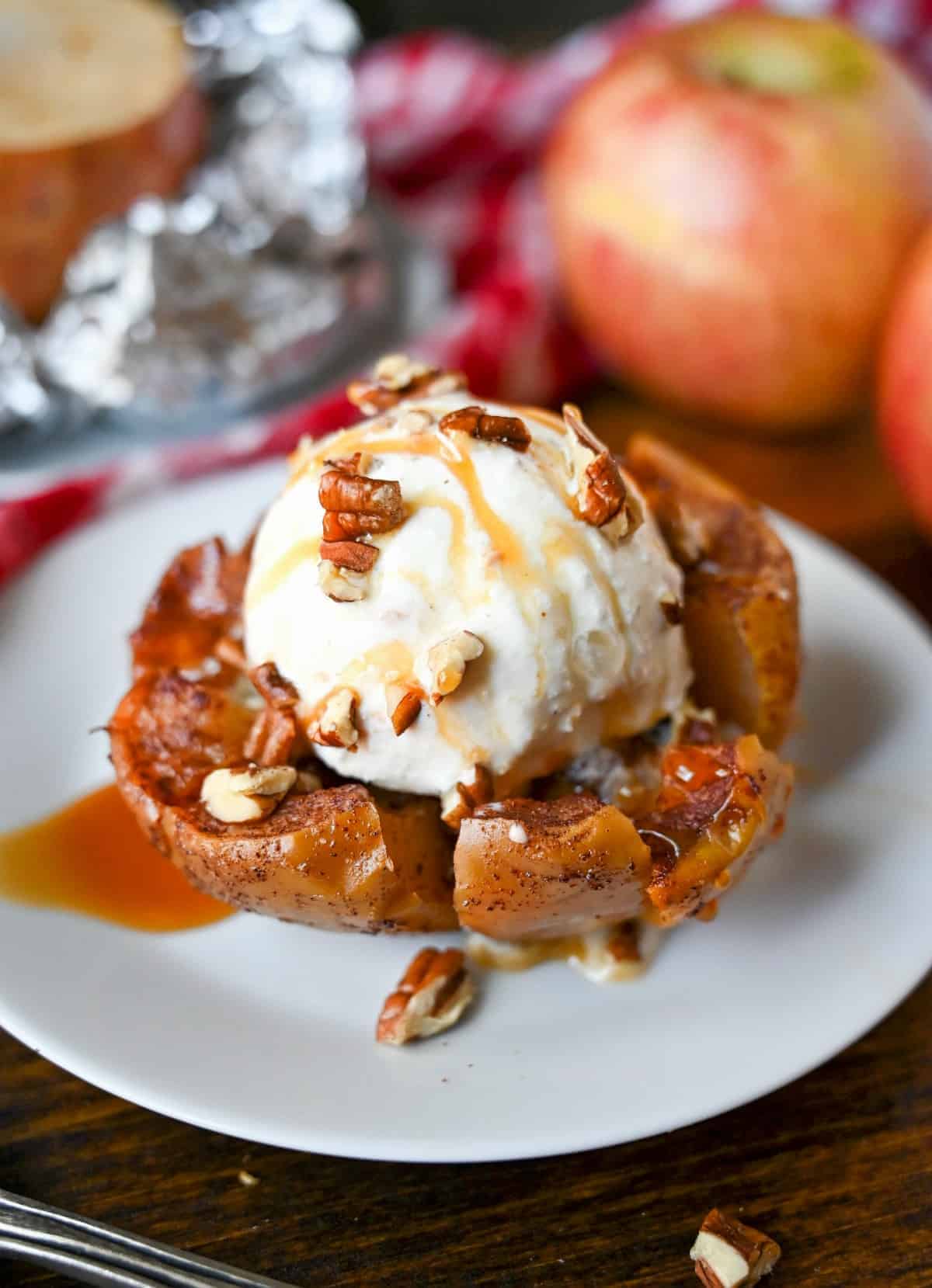 Bloomin grilled apple on a small white plate with caramel sauce and pecans on top. Two apples in the background.