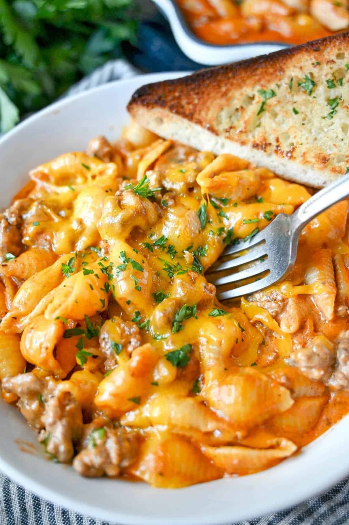 Creamy beef and pasta in a white bowl with a fork going into it and a side of garlic bread.