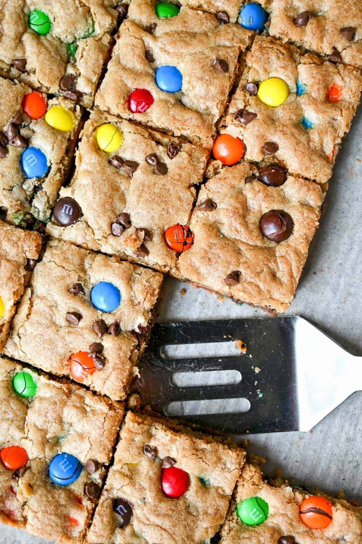 M&M cookie bars in a baking pan cut into squares and a small serving spatula.