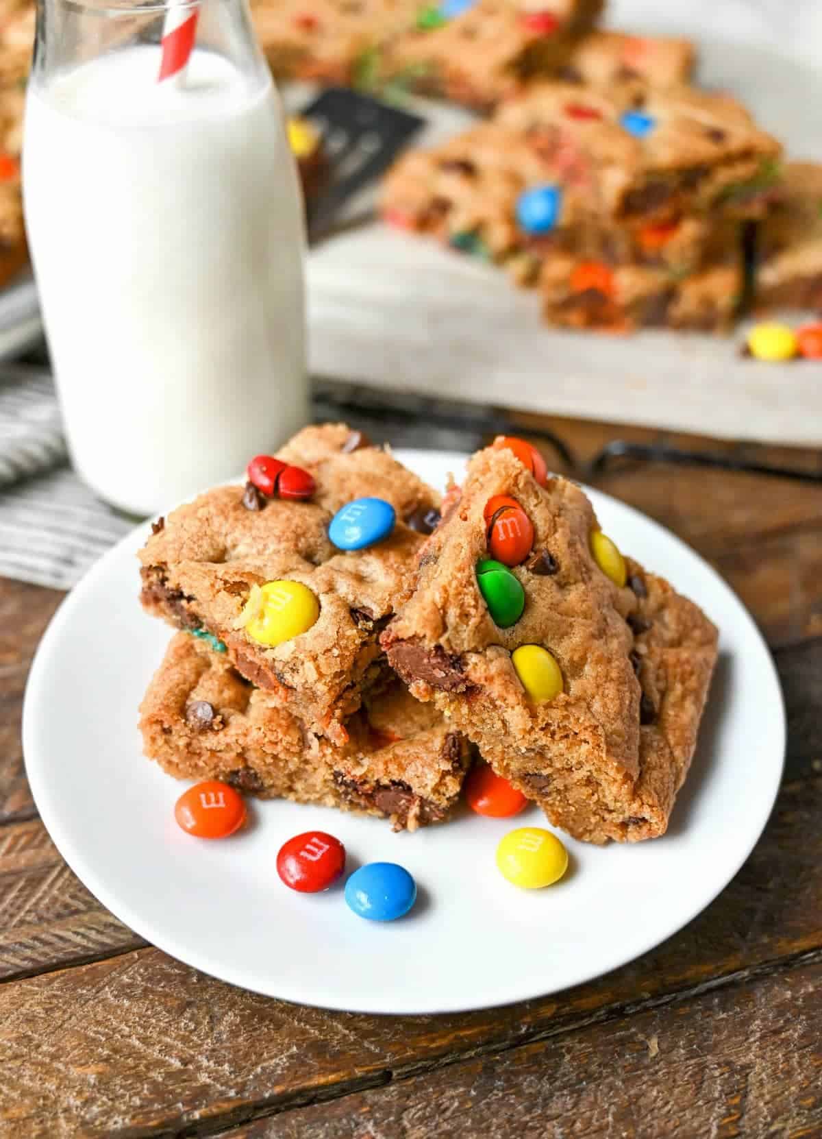 Three M&M cookie bars on a small white plate with a glass of milk.