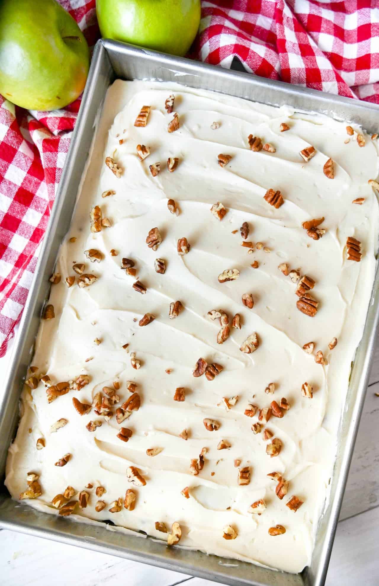 Apple cake with cream cheese frosting in a 9x13 baking pan. On a red and white checkered towel and two green apples on the side.