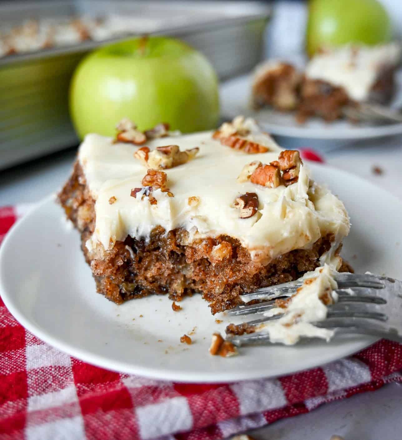 Apple cake with cream cheese frosting and pecans on top of a wgite plate and a fork on the side.