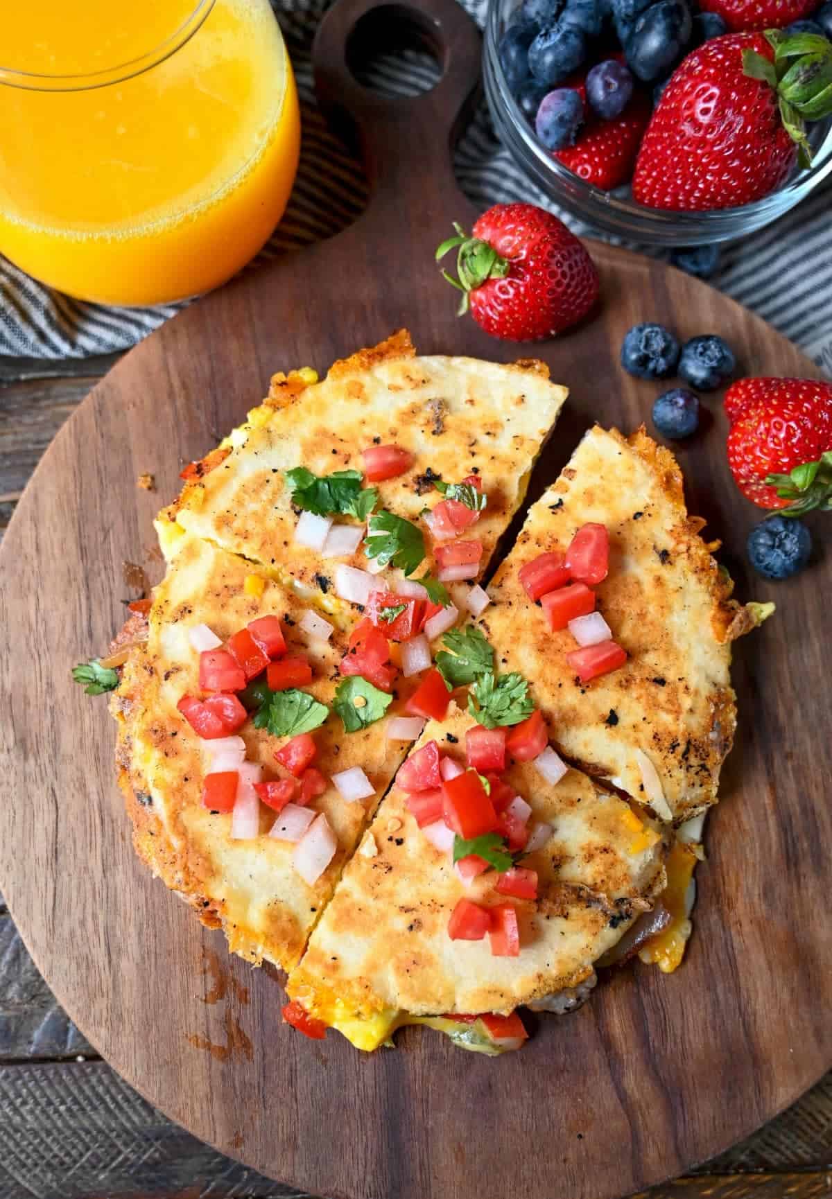 Breakfast quesadilla cut into fourths on a cutting board with a side of strawberries, blueberries and orange juice.