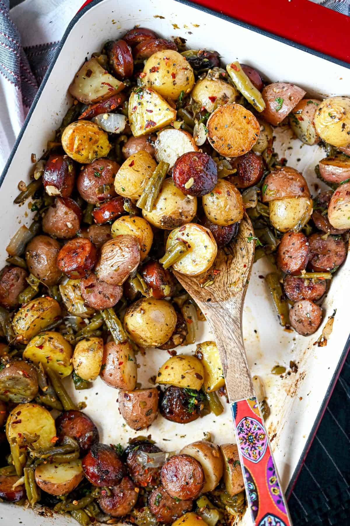 Sausage, green beans and potatoes all in a casserole dish with a wooden spoon.