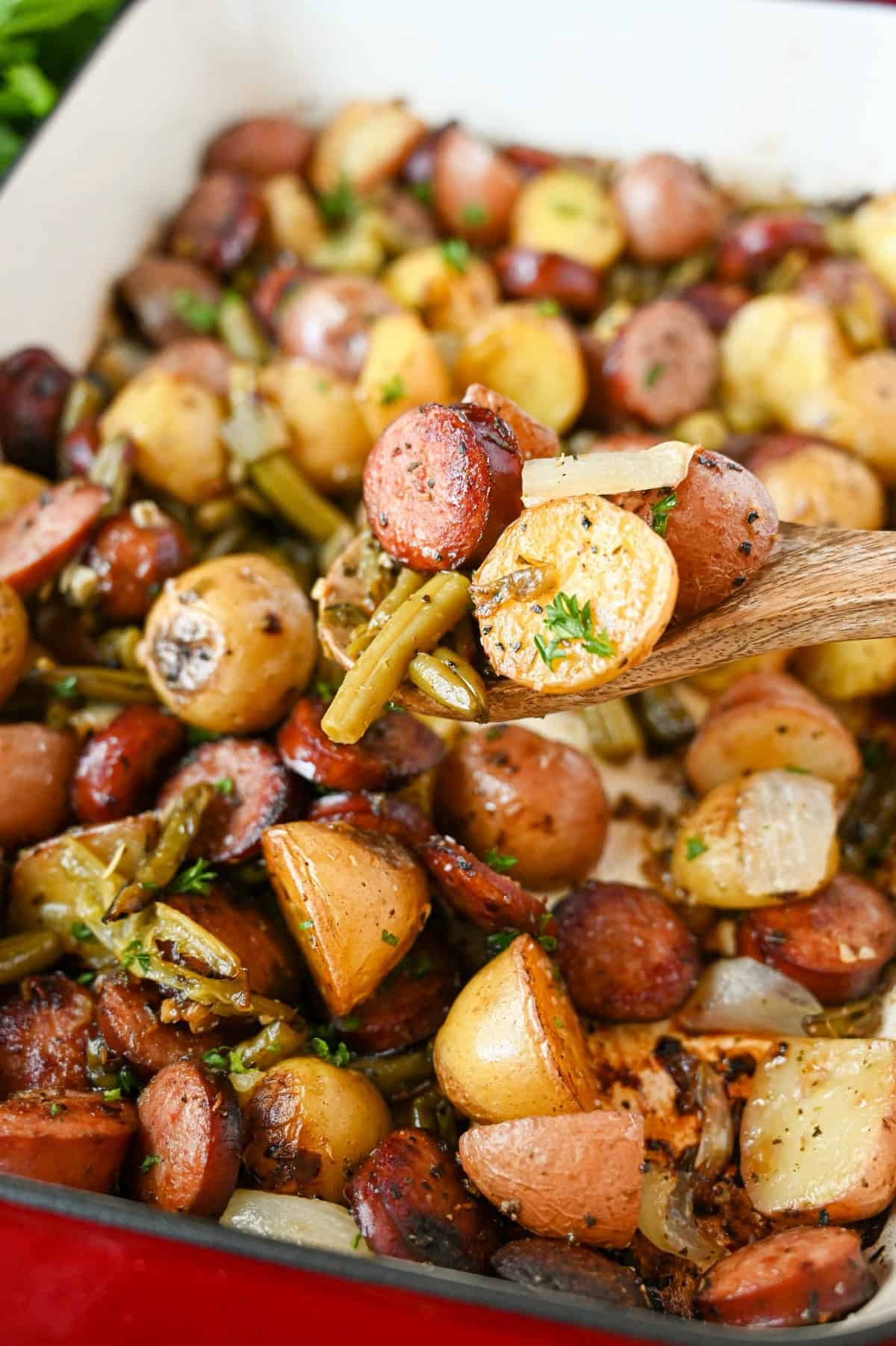 A wooden spoon scooping out some sausage, green beans and potato bake out of a casserole dish.