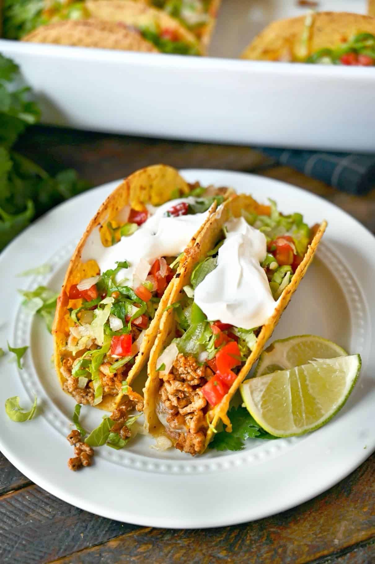 Two baked chicken tacos on a white plate with two lime slices.