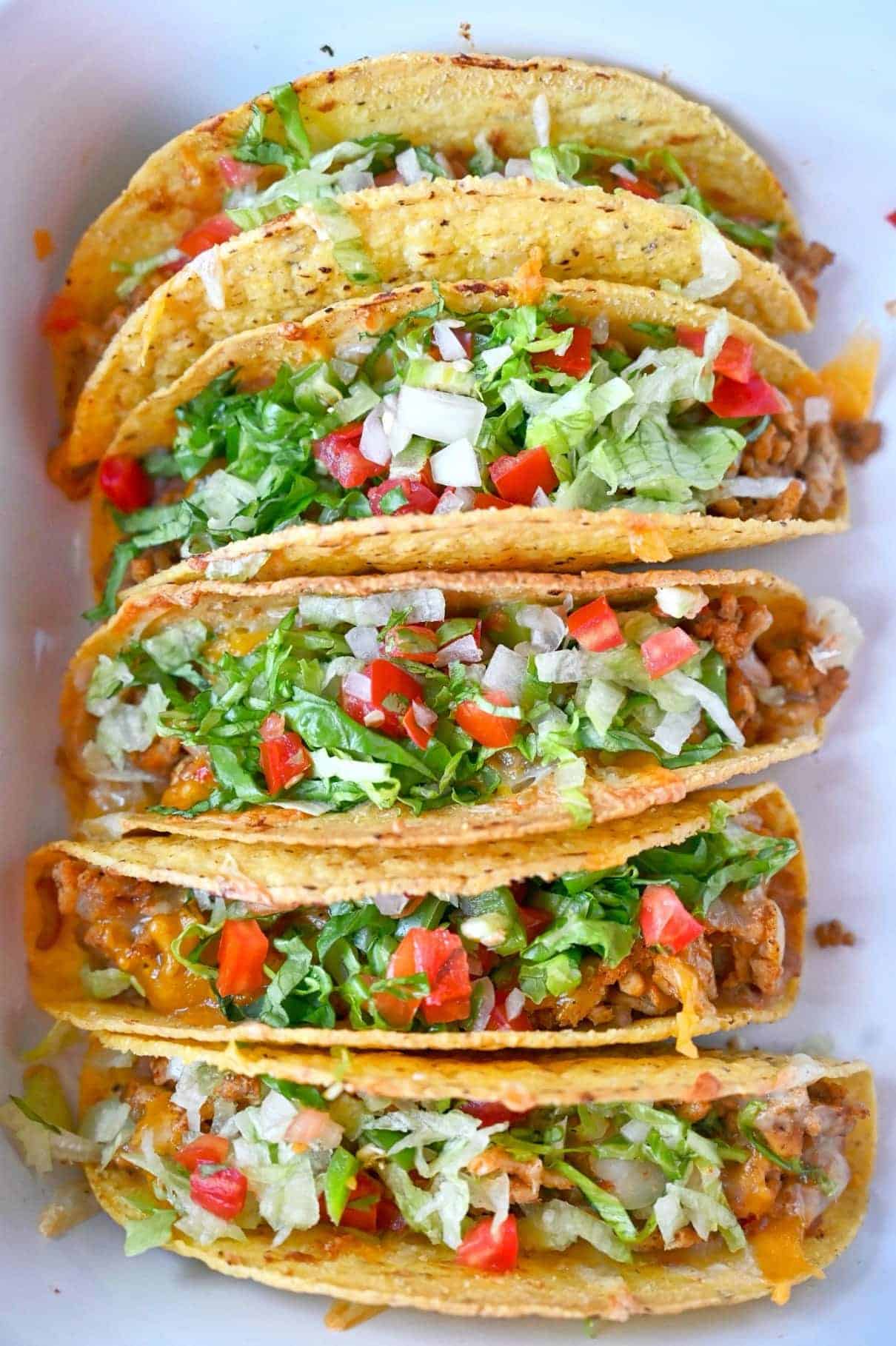 Five baked chicken tacos in a white baking dish. Topped with lettuce and pica de gallo on top.