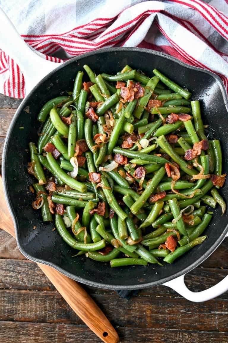 Green beans and bacon all in a skillet with a wooden spoon on the side.