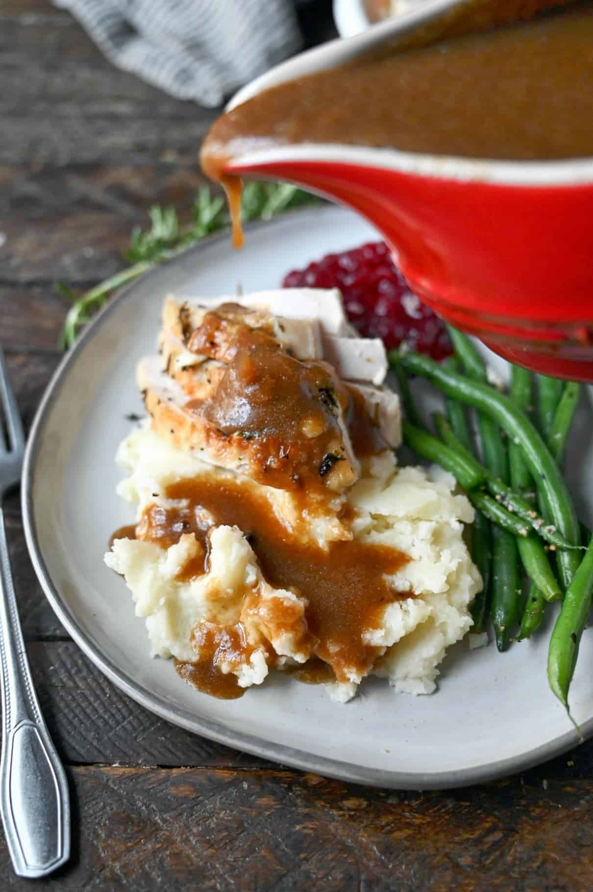 Turkey gravy being poured out of a red gravy boat on top of mashed potatoes and turkey.