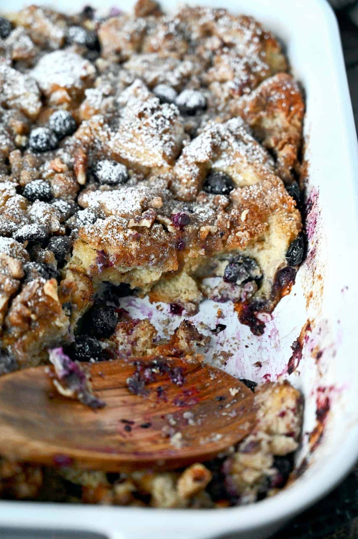 Blueberry french toast casserole in a white baking dish with a scoop missing and a wooden spoon.