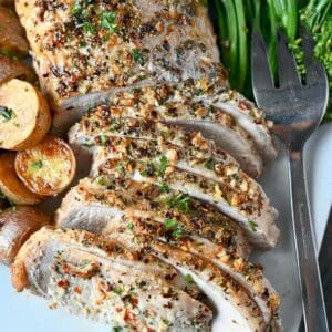 Herb crusted pork loin sliced on a white plate. With a side of broccolini and roasted potatoes.