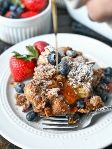 Blueberry french toast casserole on a white plate with syrup being poured on top and a side of strawberries.