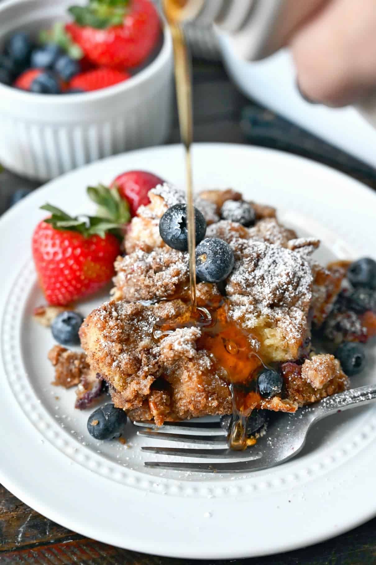 Blueberry french toast casserole on a white plate with syrup being poured on top and a side of strawberries. 