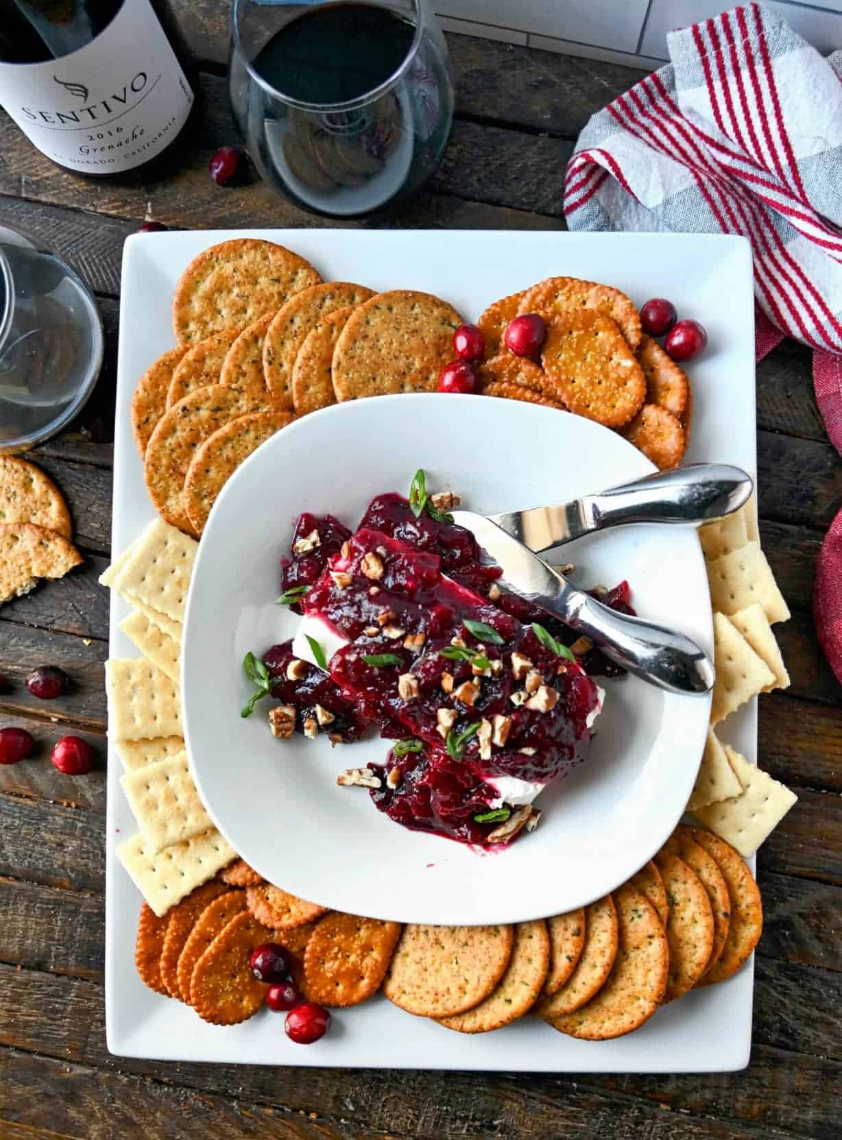 Cranberry cream cheese dip on a small white plate. placed on a white platter with crackers all around and two small serving knives.