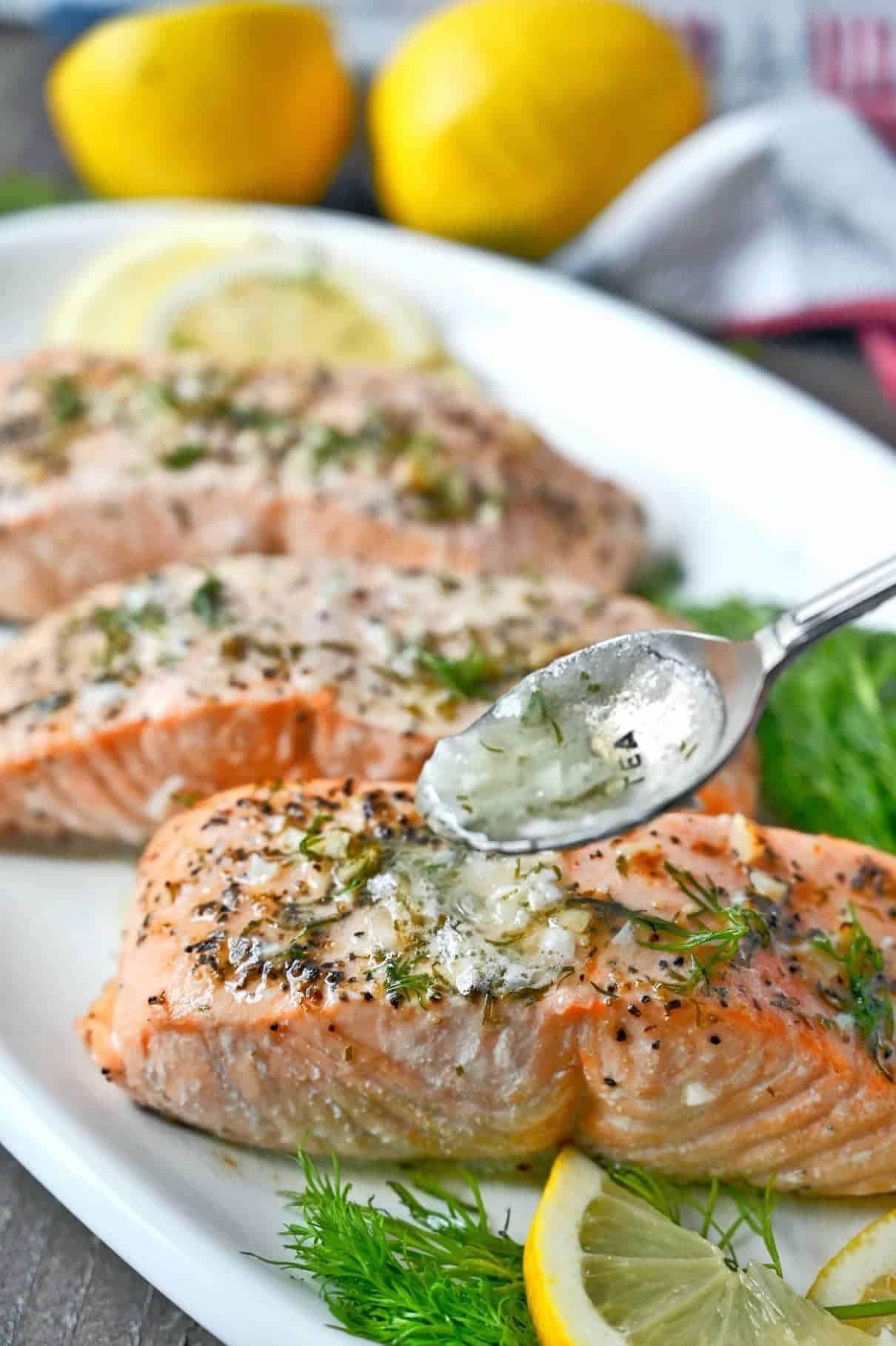 Three salmon filets on a white platter and a spoon pouring on lemon butter sauce.