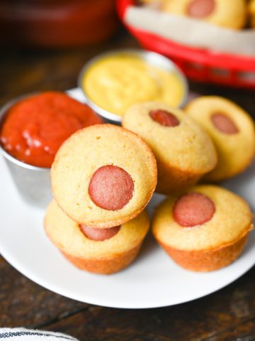A few mini corn dog muffins on a small white plate with a side of ketchup and mustard.