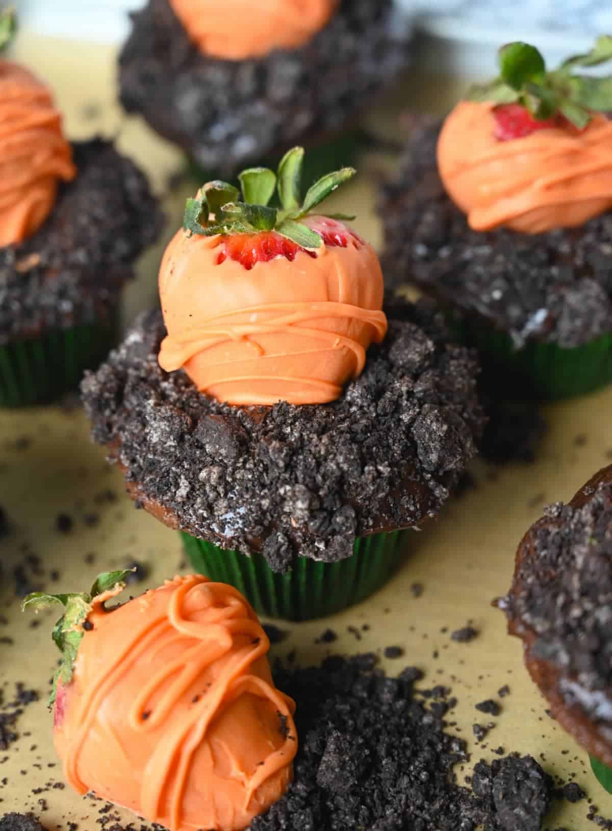 Five chocolate cupcakes with crushed oreos on top to look like dirt and a chocolate covered strawberry on top to look like a carrot.