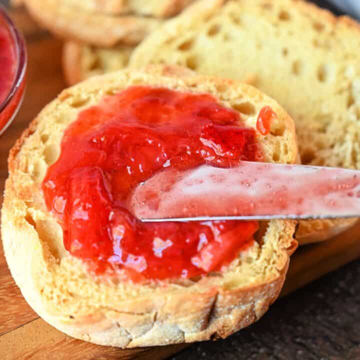 Strawberry jam being spread on an emglish muffin with a knife.
