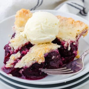 A slice of cherry pie on a white plate with a scoop of vanilla ice cream.