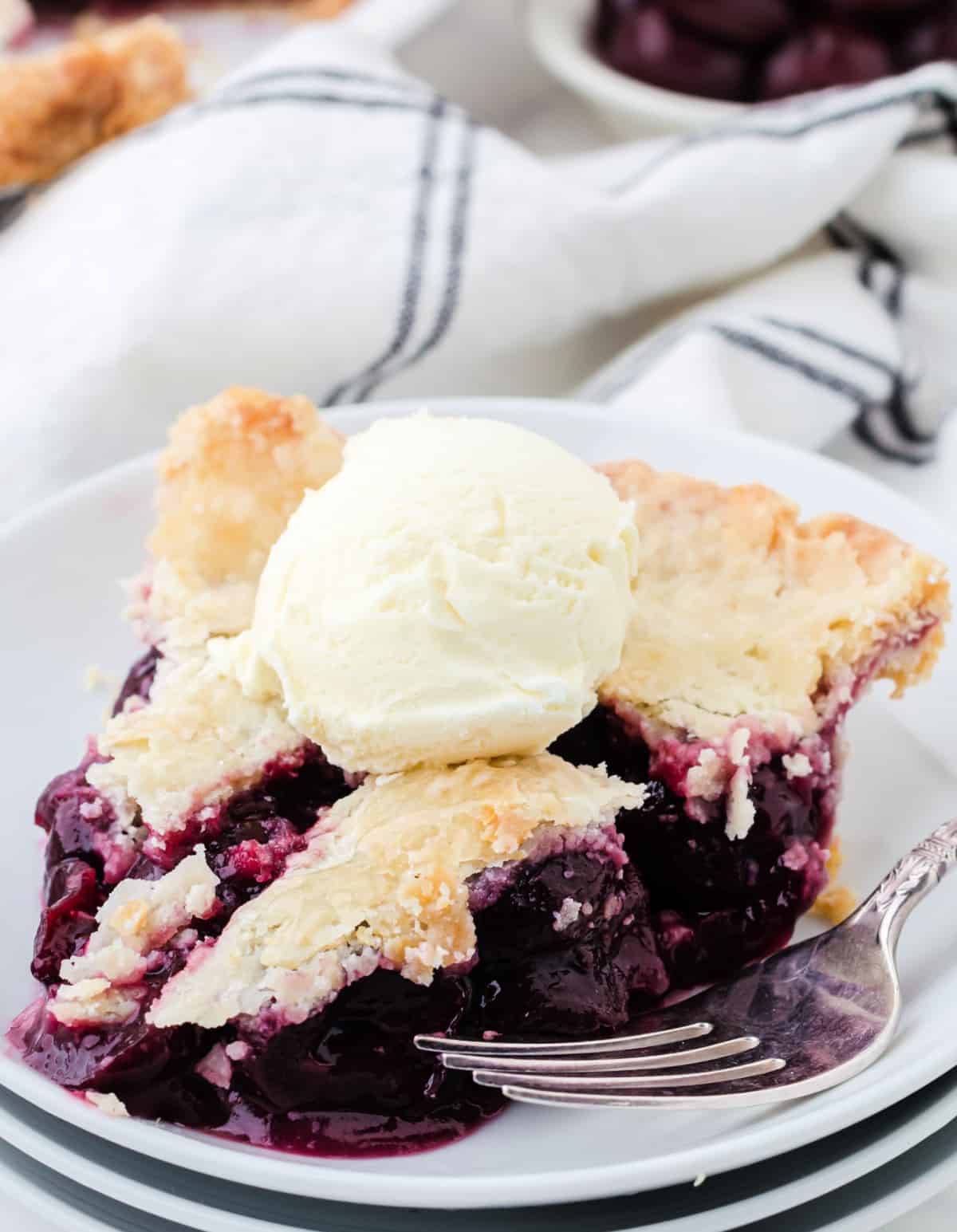 A slice of cherry pie on a white plate with a scoop of vanilla ice cream.